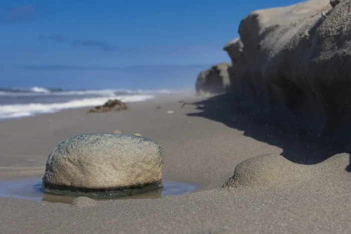 Ein runder Stein liegt am Rand einer flachen Wasserlache am Strand.