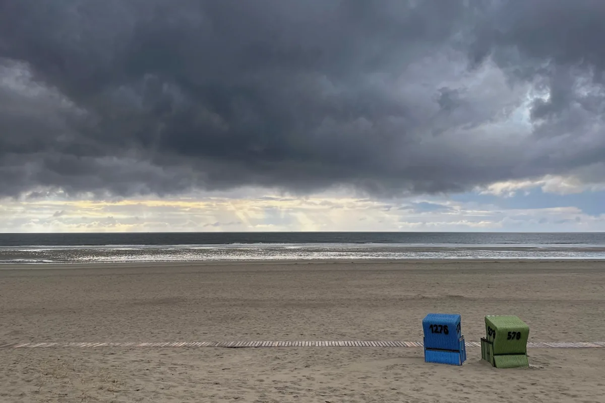 Zwei Strandkörbe, ein blauer und ein grüner, stehen am leeren Strand unter dunklen Wolken, am Horizont fällt Licht aufs Meer.